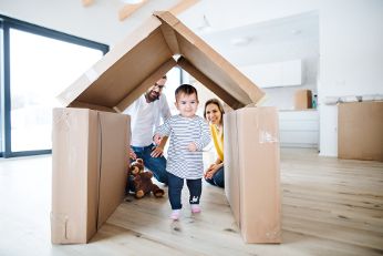 Familie mit Kind. Das Kind läuft durch ein Haus, welches aus Karton gebaut worden ist.